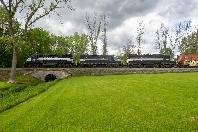 Three Finger Lakes Railway locomotives crossing a stone-arch culvert over a green lawn